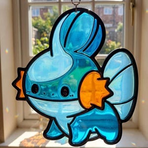 Puede incluir: Un atrapasoles de vidriera azul y naranja con un personaje de pez de dibujos animados con contornos negros. El atrapasoles cuelga de una cadena y se exhibe frente a una ventana.