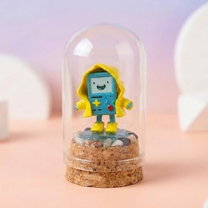 Puede incluir: Un personaje de dibujos animados azul con un impermeable y botas amarillas, dentro de una cúpula de cristal transparente. El personaje está sobre una base de pequeñas piedras y corcho. La cúpula está sobre una superficie rosa.