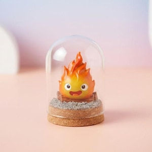 May include: A decorative figurine of a cartoon fire character with orange and yellow flames, a smiling face, and a wooden base. The figurine is encased in a clear glass dome with a cork base. The background is a soft gradient of pink and purple.