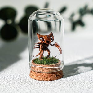 May include: A small brown creature figurine with pointed ears and spikes on its back, standing on a patch of green grass inside a clear glass cloche with a cork base. The creature has a skeletal appearance with hollow eye sockets and long limbs.