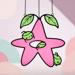 Op de afbeelding: Een roze, stervormige glas-in-lood zonnevanger met een zwarte omtrek, met drie groene cartoonfiguren en twee groene bladeren. De zonnevanger hangt aan een zwarte ketting. De achtergrond is zachtroze en wit.