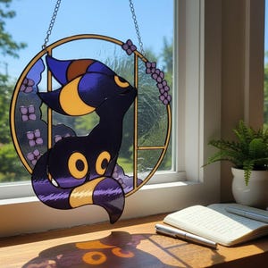 May include: Stained glass suncatcher featuring a black and purple creature with yellow accents, set within a gold-toned circular frame. The design includes purple floral elements and is suspended by a chain. The suncatcher is displayed in front of a window.