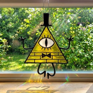 May include: A stained glass suncatcher in the shape of a yellow triangle with a single eye, a black top hat, and a black bow tie. The suncatcher has black arms and legs. It is hanging in front of a window with a green background.