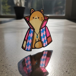 Puede incluir: Un collar con colgante que presenta un personaje de gato de dibujos animados con pelaje naranja y blanco, que lleva una capa estampada en rosa y azul. El gato tiene un peque&ntilde;o colgante azul en forma de l&aacute;grima. El collar est&aacute; suspendido por una cadena plateada.