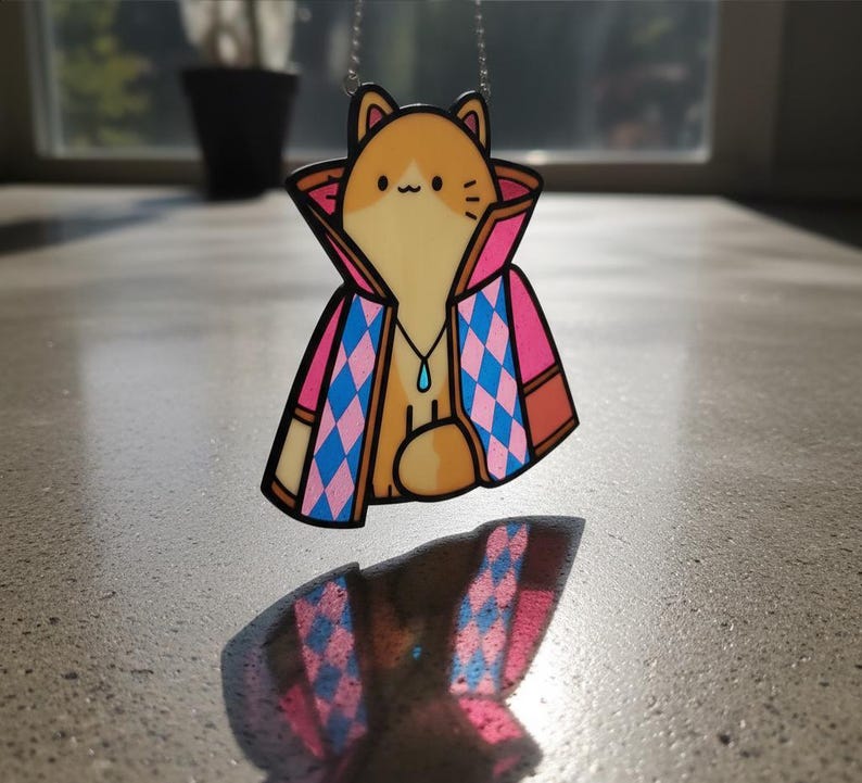 Puede incluir: Un collar con colgante que presenta un personaje de gato de dibujos animados con pelaje naranja y blanco, que lleva una capa estampada en rosa y azul. El gato tiene un peque&ntilde;o colgante azul en forma de l&aacute;grima. El collar est&aacute; suspendido por una cadena plateada.