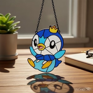 May include: A stained glass-style suncatcher featuring a cartoon penguin character with a blue and white body, yellow feet, and a gold crown. The suncatcher is suspended by a black chain and casts a shadow on a wooden surface.