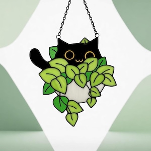 May include: A black cat suncatcher with a smiling face and gold eyes peeking out from green leaves. The cat has a black tail and is suspended from a black chain. The leaves are a bright green colour.