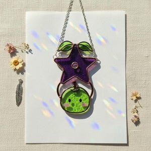 Op de afbeelding: Een gebrandschilderde zonnevanger met een paarse ster en een groene appel met een lachend gezicht. De ster heeft een koperen frame en is via een koperen ring verbonden met de appel. De zonnevanger hangt aan een zilveren ketting.