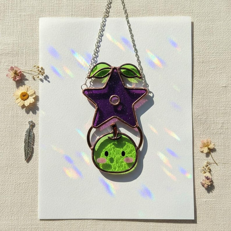 Op de afbeelding: Een gebrandschilderde zonnevanger met een paarse ster en een groene appel met een lachend gezicht. De ster heeft een koperen frame en is via een koperen ring verbonden met de appel. De zonnevanger hangt aan een zilveren ketting.