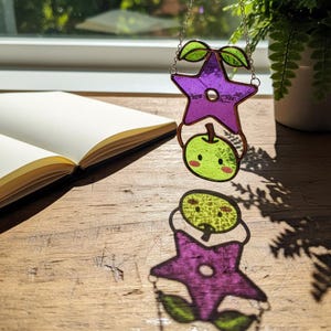 Op de afbeelding: Een gebrandschilderde zonnevanger met een paarse ster en een groene appel. De ster heeft een vijfhoekige vorm met groene bladeren. De appel heeft een schattig gezicht. Hij hangt aan een zilveren ketting.