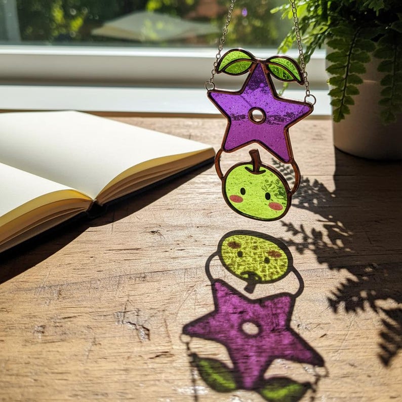 Op de afbeelding: Een gebrandschilderde zonnevanger met een paarse ster en een groene appel. De ster heeft een vijfhoekige vorm met groene bladeren. De appel heeft een schattig gezicht. Hij hangt aan een zilveren ketting.