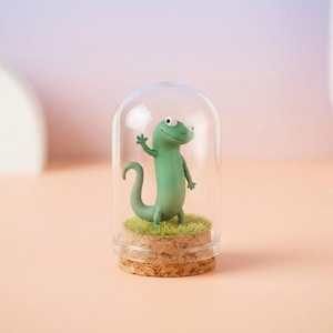 May include: A charming green lizard figure displayed under a clear glass dome. The lizard stands on a patch of artificial grass, set on a cork base. The background features a soft pink and purple gradient.