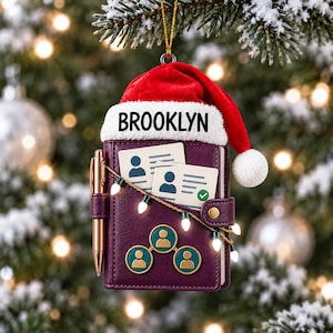 Op de afbeelding: Een paars agenda-ornament met een rood-witte kerstmuts en de tekst BROOKLYN. De agenda bevat twee ID-kaarten, een gouden pen, een snoer met kleine witte kerstlampjes en drie ronde iconen met silhouetten van personen erop afgebeeld.