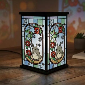 May include: A black, stained-glass lamp featuring a cartoon character with a white belly, surrounded by red flowers and green leaves. The lamp has a rectangular shape and is placed on a wooden surface.