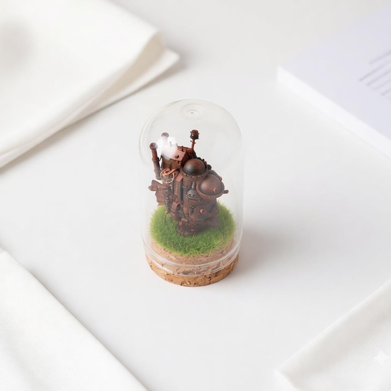 May include: A miniature, detailed brown steampunk-style castle figurine inside a clear glass dome. The base is cork, with a patch of green grass. The dome sits on a white surface, with white fabric in the background.