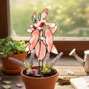 Könnte beinhalten: Ein Gartenstecker aus rosa und weißem Glas in Form einer stilisierten Blume, gepflanzt in einem Terrakottatopf mit Lavendel und Sukkulenten. Der Stecker hat rote Augen und grüne Akzente. Eine Gießkanne und andere Topfpflanzen sind im Hintergrund.