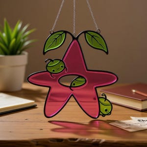 Op de afbeelding: Een roze, stervormige zonnevanger met een zwarte omtrek, met drie groene, cartoonachtige karakters. Twee groene bladeren staan bovenaan en de zonnevanger hangt aan een zilveren ketting. De zonnevanger staat op een houten oppervlak.