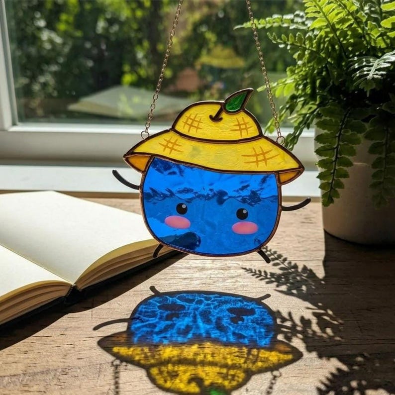 Op de afbeelding: Een gebrandschilderde zonnevanger met een grillig blauw karakter met een gele hoed en roze wangen. De zonnevanger hangt aan een delicate ketting en werpt een kleurrijke schaduw. Decoratieve kunst voor ramen.