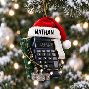 Puede incluir: Un adorno navideño que muestra una calculadora azul oscuro con un rollo de papel, un libro verde, una pluma estilográfica negra y una pila de monedas plateadas. Un gorro de Papá Noel rojo con el nombre NATHAN en el borde blanco.