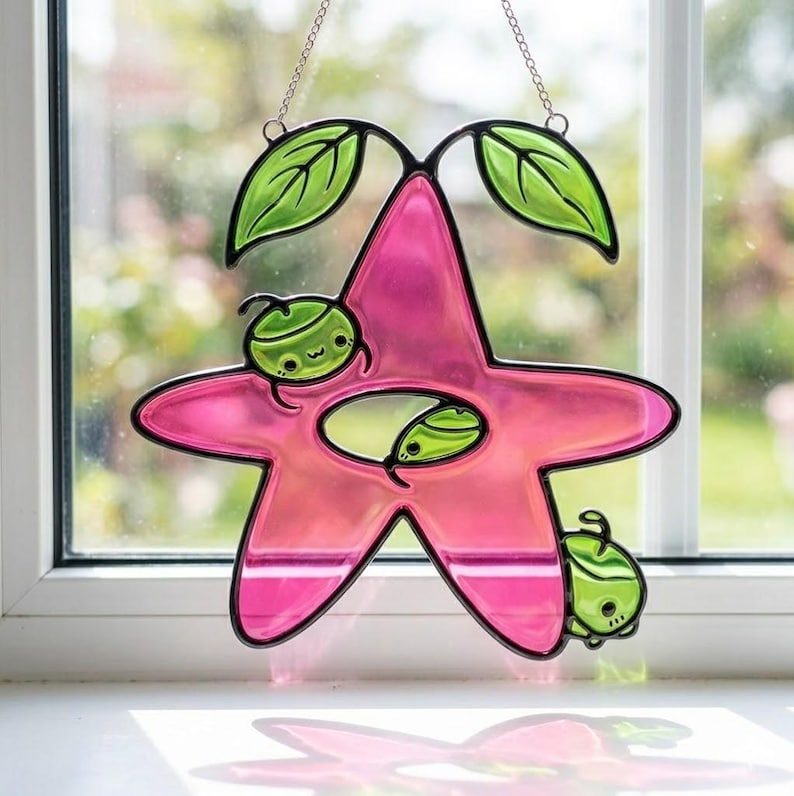 Op de afbeelding: Een roze, stervormige glas-in-lood zonnevanger met een zwarte omtrek, met groene bladeren en cartoonachtige groene vruchten. De zonnevanger hangt aan een ketting en wordt voor een raam getoond.