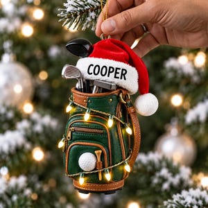 May include: A miniature green and brown golf bag ornament featuring a red Santa hat with the name COOPER printed in black. The bag contains silver golf clubs, a white golf ball, and a tee, all wrapped in a string of small glowing yellow lights.