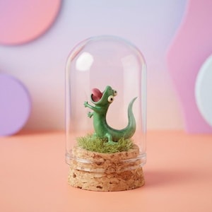 Op de afbeelding: Een grillig terrarium met een groene hagedisfiguur in een glazen koepel. De hagedis steekt zijn tong uit en zit op kunstgras, bovenop een kurken voet. De achtergrond heeft pastelkleuren.