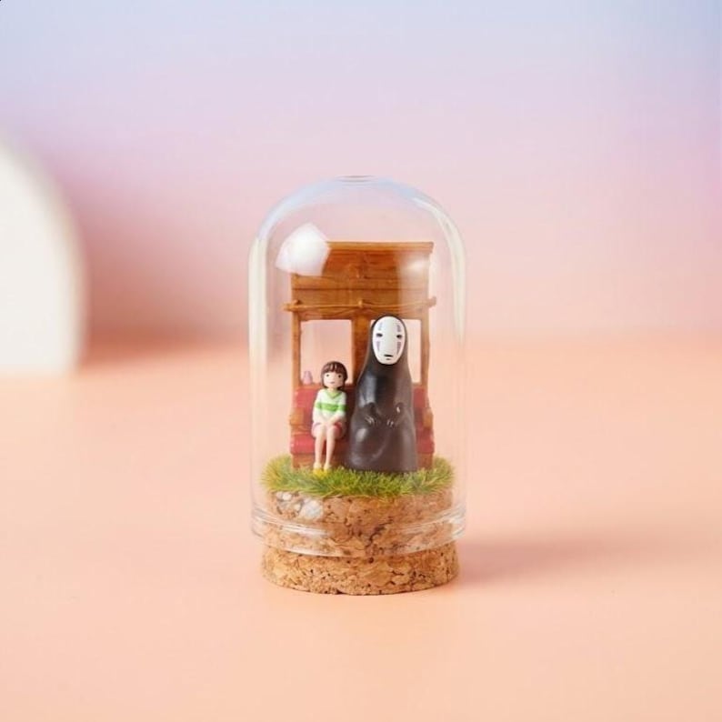 May include: A miniature scene inside a glass dome featuring figures from a popular animated film. The scene includes a wooden structure, a girl, and a tall, dark figure. The base is made of cork, and the background is a soft gradient of pink and purple.