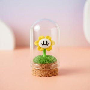 May include: A whimsical decorative piece featuring a yellow flower with a smiling face, encased in a glass dome. The flower sits atop a patch of green, textured material, and a cork base. The dome is on a pink surface.