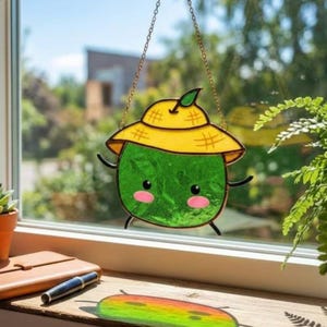Op de afbeelding: Een gebrandschilderde zonnevanger met een cartoon watermeloen karakter met een gele hoed. De groene watermeloen heeft roze wangen, zwarte ogen en kleine zwarte armen en benen. De zonnevanger hangt aan een gouden ketting.