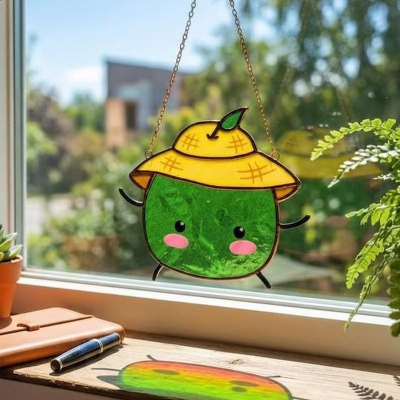 Op de afbeelding: Een gebrandschilderde zonnevanger met een cartoon watermeloen karakter met een gele hoed. De groene watermeloen heeft roze wangen, zwarte ogen en kleine zwarte armen en benen. De zonnevanger hangt aan een gouden ketting.