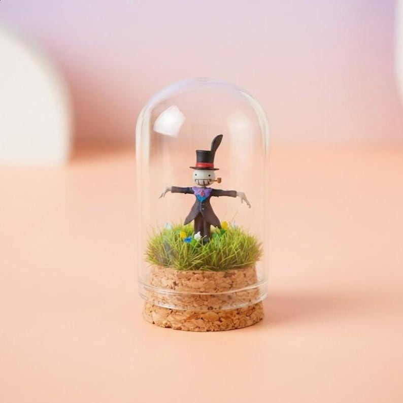 May include: A miniature figurine of a scarecrow character inside a glass dome. The figure wears a black top hat, a purple scarf, and a dark coat. It stands on a bed of green grass and small flowers, set on a cork base.