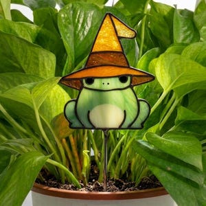 May include: A stained glass frog ornament featuring a light green body with a cream-colored belly and black eyes, wearing a pointed orange witch hat. The figure is attached to a thin metal stake, designed to be inserted into a potted plant.