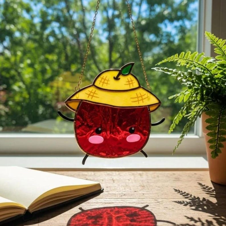 Op de afbeelding: Een gebrandschilderde zonnevanger met een rood fruitpersonage met een gele hoed en zwarte armen en benen. Het personage heeft roze wangen en hangt aan een gouden ketting. De zonnevanger wordt tentoongesteld voor een raam met een plant.