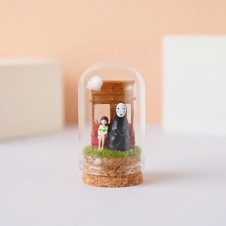 May include: A miniature glass dome terrarium featuring figures from the animated film Spirited Away. The scene includes a girl and a character in a black robe, set against a wooden structure and artificial grass, atop a cork base.