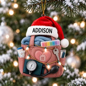 Può includere: Un ornamento a forma di borsa rosa con un cappello da Babbo Natale rosso e il nome ADDISON stampato sul bordo bianco. La borsa presenta una toppa a cuore con l'icona di una casa, uno sfigmomanometro, un portapillole, una chiave e luci natalizie.