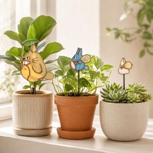 Peut inclure: Trois tuteurs pour plantes en vitrail représentant des personnages de dessin animé sur de fines tiges métalliques noires. Un grand personnage beige tient une feuille verte, un plus petit bleu a des ailes et un minuscule blanc.