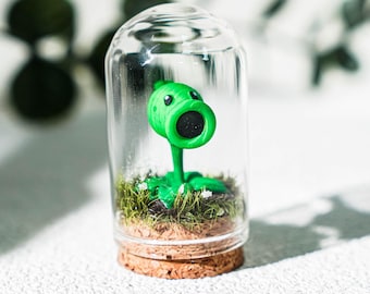 May include: A green pea shooter character figurine with a large circular mouth and dark centre, standing on a patch of green grass inside a clear glass cloche dome with a cork base. The figure has a smooth texture and a bright green colour.