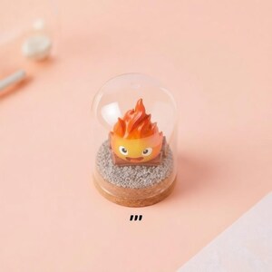May include: A miniature figurine of a fire demon, with orange and yellow flames, sits inside a clear glass dome. The base is a wooden circle with a bed of gray gravel. The figurine has a smiling face.