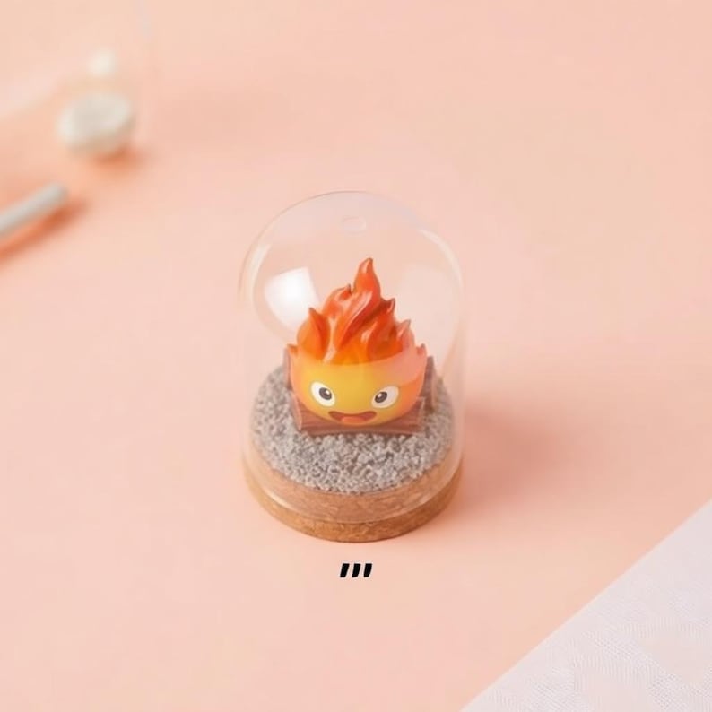 May include: A miniature figurine of a fire demon, with orange and yellow flames, sits inside a clear glass dome. The base is a wooden circle with a bed of gray gravel. The figurine has a smiling face.