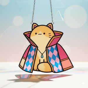 Puede incluir: Un atrapasoles caprichoso con forma de gato. El gato, de color naranja y crema, lleva una capa rosa y azul estampada con un colgante turquesa. Cuelga de una cadena plateada, ideal para decorar ventanas o paredes.