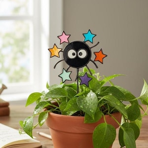 May include: A black circular creature ornament with two large white eyes, featuring six colorful stars in pink, blue, orange, light green, and purple attached to its perimeter, mounted on a thin metal stake inside a terracotta plant pot.