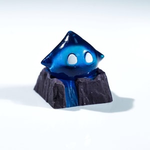 May include: A blue and black artisan keycap depicting a stylized creature with white eyes and a pointed head. The keycap sits on a rocky base with a blue waterfall design. Designed for mechanical keyboards.