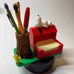 May include: A Snoopy-themed desk organizer featuring a pen holder shaped like a tree trunk, a red doghouse with a Snoopy figurine, and a yellow Woodstock figurine. The organizer includes pens, pencils, and sticky notes.