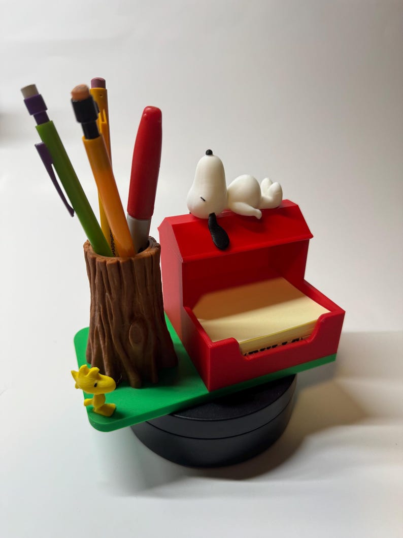 May include: A Snoopy-themed desk organizer featuring a pen holder shaped like a tree trunk, a red doghouse with a Snoopy figurine, and a yellow Woodstock figurine. The organizer includes pens, pencils, and sticky notes.