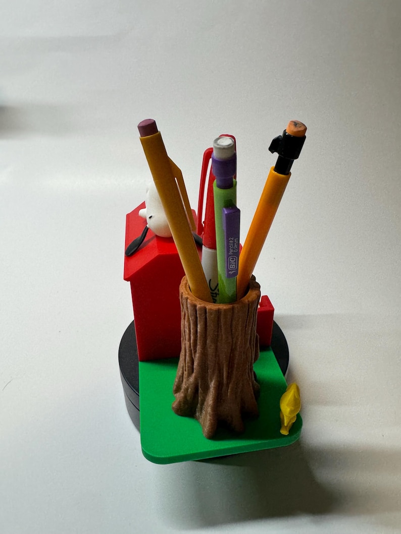 May include: A novelty desk organizer featuring a brown tree trunk pen holder, a red dog house, and a green base. The organizer holds several pens and pencils. A yellow bird figurine is on the green base. A white dog figurine is on the red dog house.