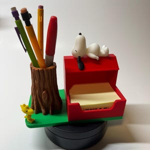 May include: A desk organizer featuring a Snoopy figurine lying on a red doghouse, with a tree trunk pen holder and a yellow Woodstock figure. The organizer includes a notepad and several pens.