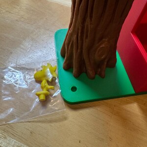 May include: A yellow cartoon character figurine and a brown tree trunk-shaped container sit on a green base. The tree trunk has a hollow center. The figurine is in a clear plastic bag. The base is on a wooden surface.