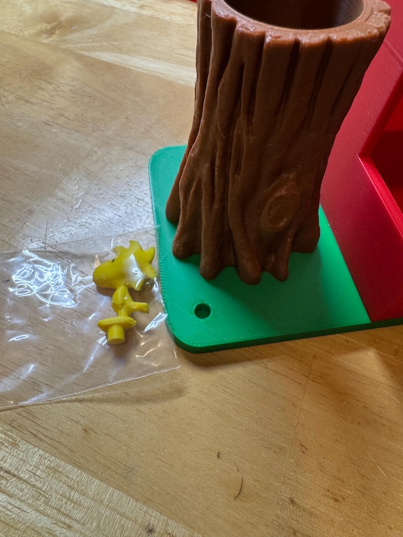 May include: A yellow cartoon character figurine and a brown tree trunk-shaped container sit on a green base. The tree trunk has a hollow center. The figurine is in a clear plastic bag. The base is on a wooden surface.