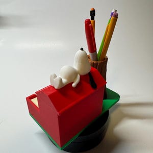 May include: A Snoopy-themed desk organizer featuring a red doghouse with Snoopy resting on top. The organizer includes a pen holder with pens and pencils. The base is black, and the doghouse has green trim.