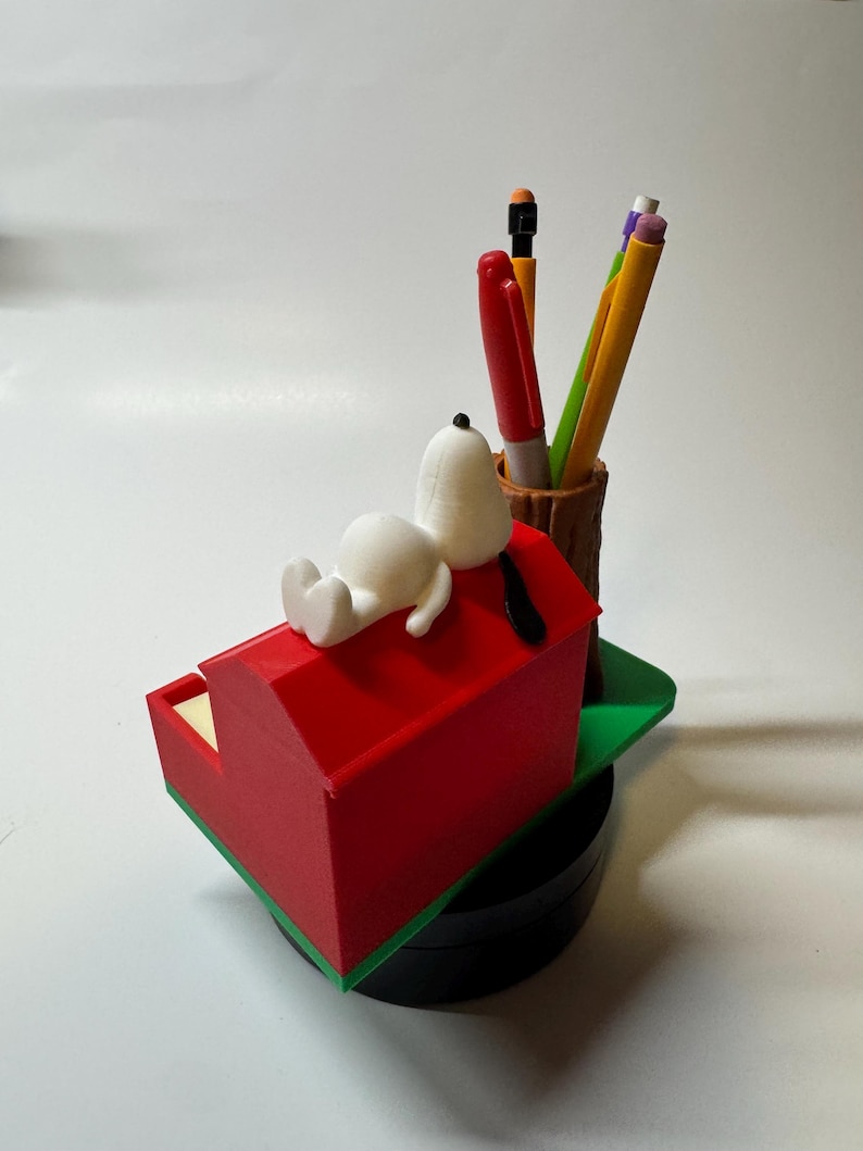 May include: A Snoopy-themed desk organizer featuring a red doghouse with Snoopy resting on top. The organizer includes a pen holder with pens and pencils. The base is black, and the doghouse has green trim.